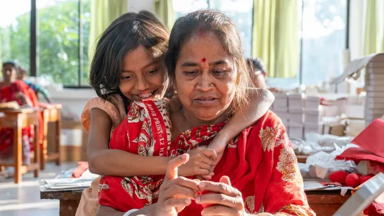 A woman sits working at a table while a young person embraces them from behind.