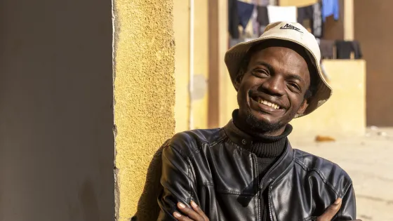 + A person in a leather jacket and hat stands by a wall, smiling. In the background, there are balconies with clothes hanging.