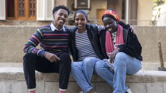 Three individuals sit closely on a stone bench, smiling and posing together. They wear casual clothing and appear relaxed.
