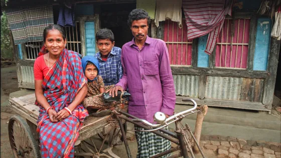  A woman, man and two children pose on a cart in front of a wooden house with barred windows and hanging cloth.