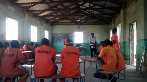 Prison inmates sit at tables listening to someone speak