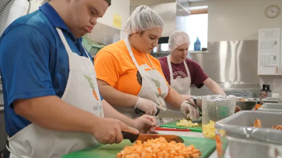 Three cooks chop vegetables at a countertop in an industrial kitchen