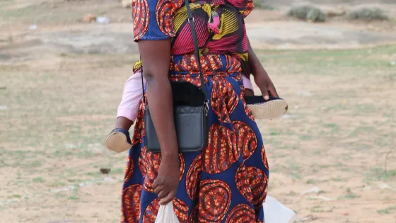 woman holding baby on back with bags of ground corn and beans at her feet