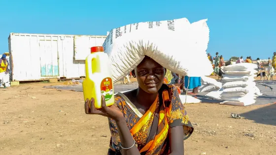 A woman from South Sudan named Martha squats in an open area next to a food distribution. She's holding food she recently received.