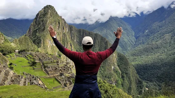 a man stands facing away from the camera with hands raised to the sky in front of Machu Picchu