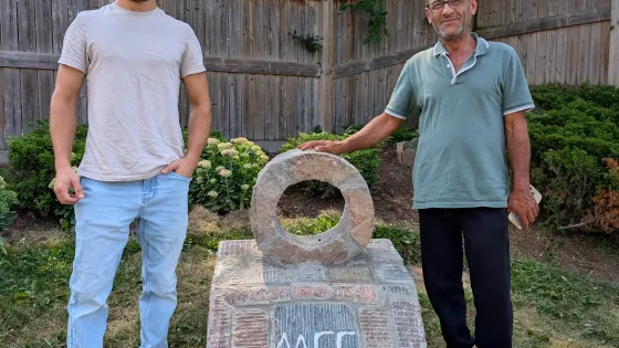 A young man and a middle-aged man stand next to a stone monument with the words MCC carved into it. The older man’s hand is resting on a stone circle on top of the monument.