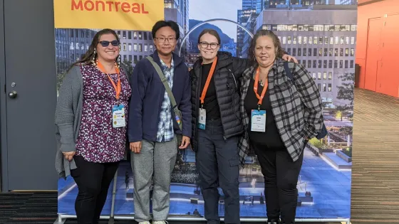Three women and a man stand in front of a banner with the words Montreal on it, smiling at the camera.