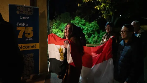A smiling group carrying various international flags walk into a building with a welcome sign that reads, "IVEP at 75 years."