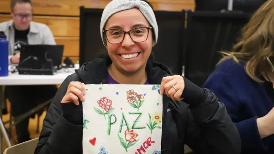 A smiling woman with glasses and dark hair holds up a square piece of cloth with the words "Paz" and "amor" on it