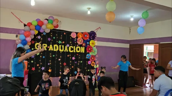 A decorated room with balloons, a "Graduación" banner, children performing, and adults watching from seated positions.