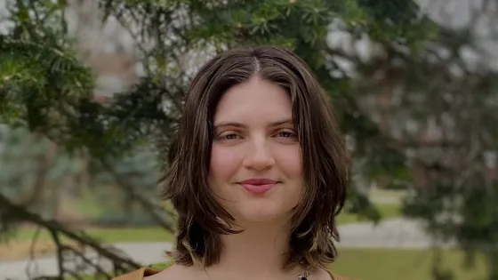 A young person with medium-length hair stands in front of a tree, wearing a fitted top and a necklace, smiling softly.