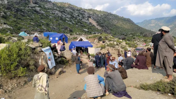  The photograph shows a mountainous landscape with tents, people gathered, and rocky terrain. Some individuals are seated or standing.