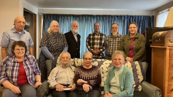 + A group of twelve people poses together in a living room, seated and standing around a couch with various furnishings in the background.