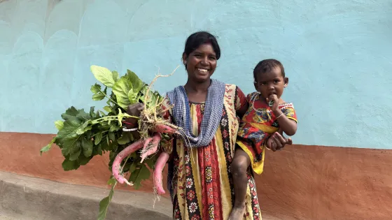 A woman from India holds her child and radishes in front of a wall. 