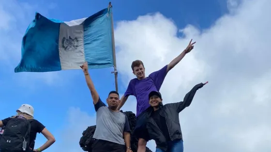 Three individuals stand on a rocky surface, one holding a flag, while others are in the background. The scene is outdoors with a mountainous backdrop.