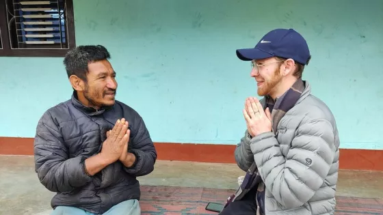 Two people sit facing each other, mirroring body language of hands pressed together in a Nepalese greeting.