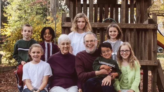 A group of two adults and seven children pose together, smiling on a wooden play structure in an outdoor setting with trees in the background.