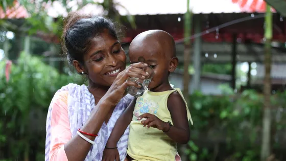 Minati Kerketta helps her son Manab Oraon drink from a cup full of clean water drawn from her Little Flock Fellowship provided water filter.