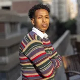 A person stands with arms crossed on a balcony, overlooking a cityscape. There are wooden chairs and tables in the background.